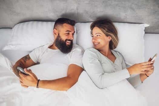 husband and wife enjoying a carefree moment lying in bed together while smiling and chatting on their smartphone in a warm bedroom setting photo